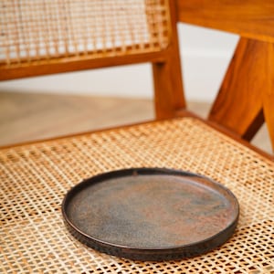 Puede incluir: Un plato redondo y oscuro con una superficie texturizada descansa sobre una silla de rat&aacute;n tejido. El plato tiene un borde ligeramente elevado y un aspecto r&uacute;stico y envejecido. La silla es de madera con un acabado natural.