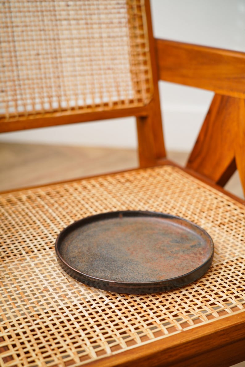 Puede incluir: Un plato redondo y oscuro con una superficie texturizada descansa sobre una silla de rat&aacute;n tejido. El plato tiene un borde ligeramente elevado y un aspecto r&uacute;stico y envejecido. La silla es de madera con un acabado natural.