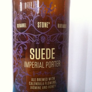 May include: A brown glass bottle with a purple label featuring the text "STONE SUEDE IMPERIAL PORTER." The label also includes "ALE BREWED WITH CALENDULA FLOWERS, JASMINE AND HONEY." The bottle contains 1 pint, 6 fl oz of ale.