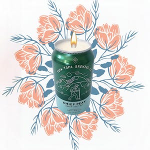 May include: A green Topa Topa Brewing Co. candle in a can, with the text "Chief Peak India Pale Ale." The candle has a lit wick and is surrounded by a floral design with peach and blue accents. The candle is a unique home decor item.