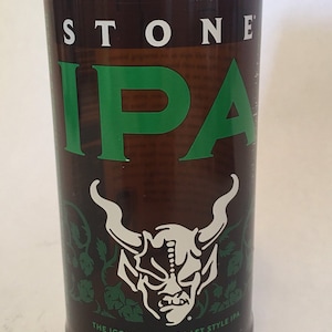 May include: A brown glass bottle with a cork stopper, featuring the text "STONE IPA" in green. The bottle has a white and green graphic of a horned demon and the text "THE ICONIC WEST COAST STYLE IPA".