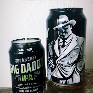May include: Two black beer can candles. One smaller can features the text "SPEAKEASY BIG DADDY IPA" with a description of the flavor. The larger can has a black and white illustration of a man in a suit and hat.