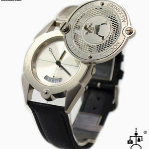 May include: A silver wristwatch with a black leather band. The watch face is white with black hands and numbers. The watch has a unique design with a circular cover that can be flipped open to reveal a silver design with the text "HI-TEK DESIGNS LONDON ALEXANDER".