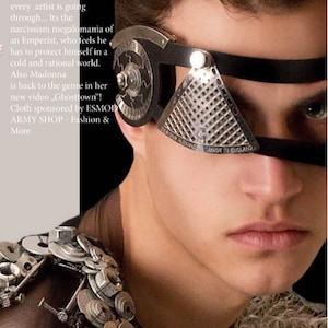 Op de afbeelding: Een close-up van een persoon die een zwart en zilveren gezichtsmasker draagt met een driehoekig ontwerp. Het masker heeft een metalen gaas en de tekst "ONS ALEXANDMADE IN ENGLAND".