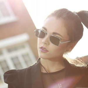 Op de afbeelding: Een vrouw draagt een zwart blazer met een zilveren ketting en een zonnebril. De zonnebril heeft een zilveren montuur en donkere glazen. De vrouw kijkt naar de zijkant.