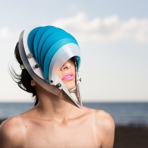 Puede incluir: Una mujer lleva una máscara facial azul y blanca con un marco de metal plateado. La máscara está hecha de paneles curvos superpuestos. La mujer mira hacia arriba y lleva lápiz labial rosa. El fondo es una imagen borrosa de una playa y el cielo.