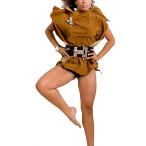 May include: A woman wearing a brown suede top with a silver buckle belt, posing with one leg raised and her hair blowing in the wind.
