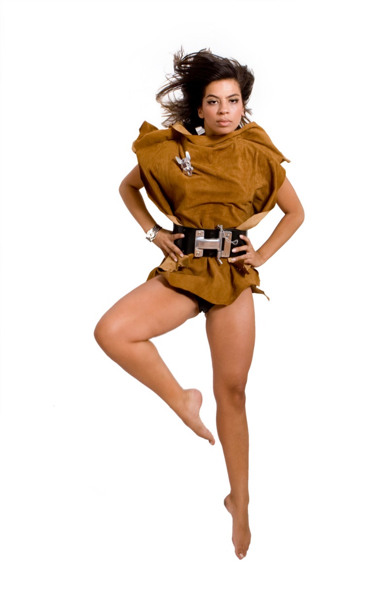 May include: A woman wearing a brown suede top with a silver buckle belt, posing with one leg raised and her hair blowing in the wind.
