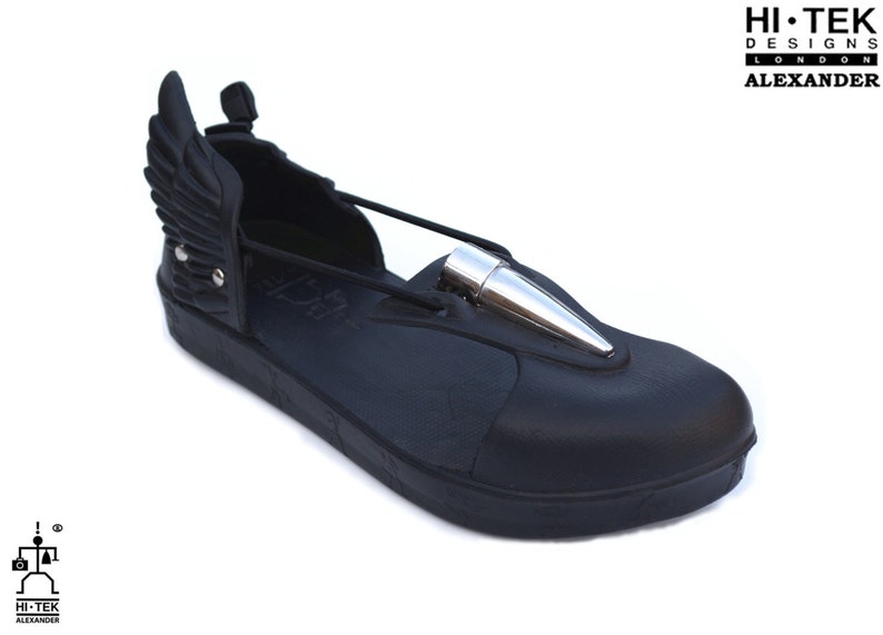May include: Black leather sandal with a silver bullet detail and a wing design on the back. The sandal has a thick black sole and a black strap across the front. The brand name "HI-TEK DESIGNS LONDON ALEXANDER" is printed on the sandal.