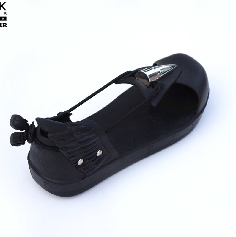 May include: Black leather sandals with a silver wing detail and a silver spike on the strap. The sandals have a black rubber sole.