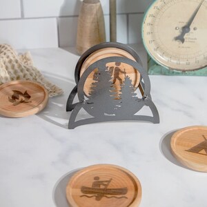 May include: Set of four wooden coasters with different nature-themed designs, including a hiker in the woods, a campfire, a canoe, and a tent. The coasters are displayed in a black metal stand.