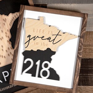 May include: Two wooden signs in the shape of the state of Minnesota. The sign on the left has a black background with a wood cutout of the state with a pattern of trees. The sign on the right has a white background with a wood cutout of the state. The top half of the state is wood and says "Life is great in the" and the bottom half is black with white numbers "218".