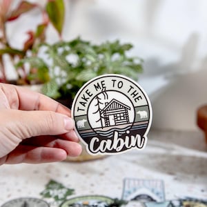 May include: A round sticker with the text "TAKE ME TO THE CABIN" encircling a cabin illustration. The cabin is surrounded by trees and bears. The sticker is held by a hand, with other stickers visible in the background.