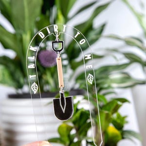 May include: Clear acrylic garden stake with the text "TALK DIRT TO ME" in white. A small shovel charm hangs from the top. The stake is designed to be placed in a potted plant.