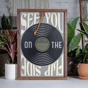 May include: A wooden framed sign with a vinyl record design. The sign reads "See You On The Flip Side" in a retro font. The record has a brown leather needle arm.