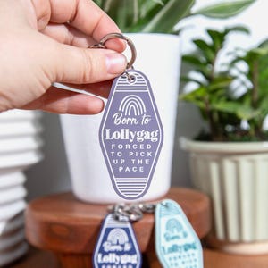May include: A purple motel-style keychain with the text "Born to Lollygag, Forced to Pick Up the Pace." The keychain has a rainbow graphic and a silver keyring. Other keychains in blue and teal are also visible.