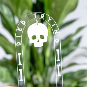 May include: Clear acrylic plant stake with the words "Died Trying" and a white skull charm. The stake has a curved top and bone-shaped accents. The background is a blurred view of green plants.