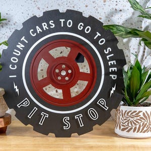 May include: A circular black wall sign shaped like a tire with a red wheel center. The text reads I COUNT CARS TO GO TO SLEEP and PIT STOP, accented by two white lightning bolt symbols. The design features a white circular border.