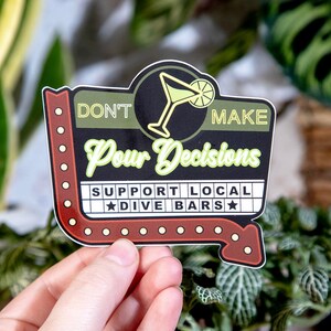 May include: A sticker with a retro sign design. The sticker features a cocktail glass illustration and the text "DON'T MAKE Pour Decisions SUPPORT LOCAL DIVE BARS" in a mix of fonts and colors, including green, black, and red.