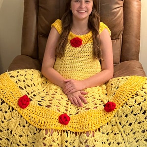 May include: A young person wearing a yellow crocheted dress with red rose embellishments. The dress is long and flowing, and the person is sitting in a brown leather armchair.