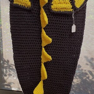 May include: A gray crocheted blanket with a yellow dinosaur tail and two yellow dinosaur pockets. The blanket is being held by a person wearing white shoes.