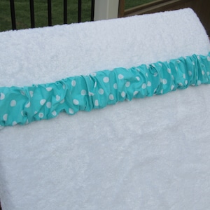 May include: White terry cloth towel with a turquoise and white polka dot band. The band is gathered and sewn onto the towel. The towel is on a wooden surface.