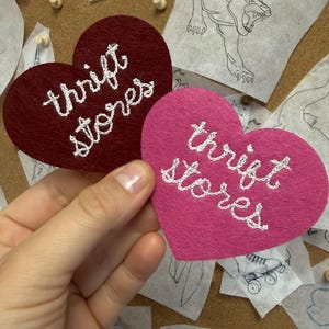 May include: Two heart-shaped felt patches, one in deep burgundy and one in bright pink, each featuring the words "thrift stores" embroidered in white thread. The patches are held up against a cork board covered in various black and white sketches.
