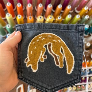 May include: A dark gray denim pocket featuring an embroidered design of a golden-brown fox in a curled position. The fox is outlined in white thread with small white dashes across its back, showcasing detailed stitching on the fabric surface.
