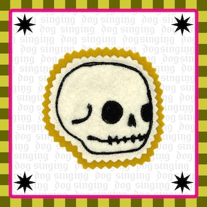 May include: A circular felt patch featuring a white skull with black embroidered details, surrounded by a mustard yellow scalloped border. The background is white with repeating light gray text reading singing dog and four black stars.