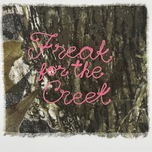 May include: A rectangular fabric patch featuring a camouflage pattern in shades of brown, green, and black. The text "Treat for the Creek" is embroidered in bright pink thread across the center in a cursive script with frayed edges.