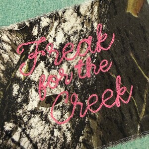 May include: A rectangular fabric patch featuring a camouflage pattern in shades of brown, black, and white. The text "Freak for the creek" is embroidered across the center in bright pink thread using a cursive, handwritten-style font.