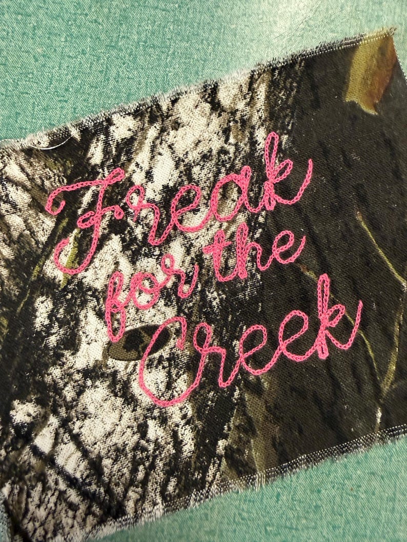 May include: A rectangular fabric patch featuring a camouflage pattern in shades of brown, black, and white. The text "Freak for the creek" is embroidered across the center in bright pink thread using a cursive, handwritten-style font.