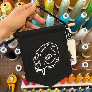 May include: Black felt banner with a white embroidered skull design. The banner is held by a black cord and a wooden dowel. The background features spools of colorful thread.