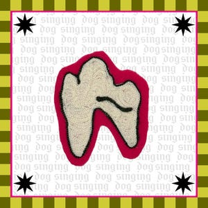 May include: A tooth-shaped textile art piece with a white center, outlined in black, and a red border. The background features the repeated text "singing dog" in a light grey color, with black starbursts in each corner. The artwork is framed with a green and yellow striped border.