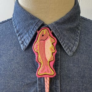 May include: A bolo tie featuring a fish-shaped pendant with pink, light pink, and yellow embroidery. The pendant is outlined in bright pink and attached to a braided light pink cord, worn over a dark blue denim shirt on a mannequin torso.