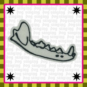May include: A light gray felt patch shaped like a canine jawbone with dark gray embroidered outlines. The background features a repeating pattern of the text "singing dog" in light gray, four black stars, and a yellow and green checkered border.