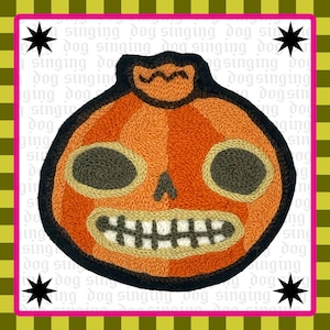 May include: An embroidered patch featuring a stylized orange pumpkin with a black stem, dark gray oval eyes, a small triangular nose, and a wide mouth with white teeth and black stitching, all outlined in a thick black border on a white background.