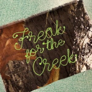 May include: A rectangular fabric patch featuring a brown, black, and tan camouflage pattern with the text "Freak for the Creek" embroidered in bright lime green thread. The edges of the patch are frayed, and the Mossy Oak logo is visible.