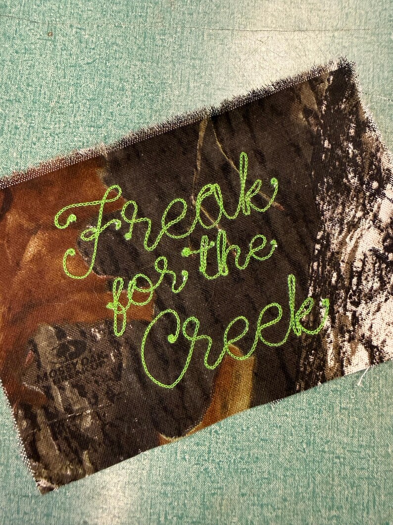 May include: A rectangular fabric patch featuring a brown, black, and tan camouflage pattern with the text "Freak for the Creek" embroidered in bright lime green thread. The edges of the patch are frayed, and the Mossy Oak logo is visible.