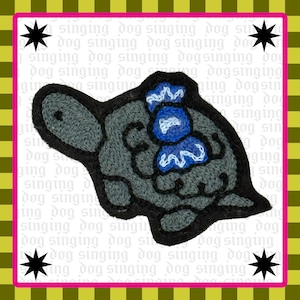 May include: A dark gray embroidered patch shaped like a turtle with a black border. The turtle shell features a bright blue wrapped candy graphic with white stitched details. The background is white with repeating text reading singing dog.