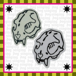 May include: Two patches featuring a stylized dog skull design with a heart detail, outlined in black on a light background. The patches are set against a patterned background with the text "singing dog" and starburst accents.