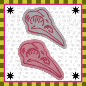 May include: Two bird skull patches, one light gray and one pink, with pink embroidered outlines and details. The background features a repeating pattern of the words "singing dog" and starburst shapes.