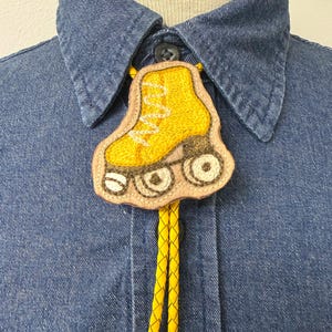 May include: A bolo tie featuring a yellow embroidered roller skate pendant with brown wheels, attached to a braided yellow cord. The pendant is mounted on a light tan leather backing and is worn over a blue denim button-down shirt.