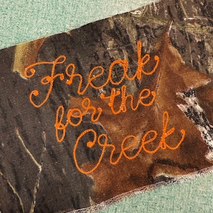 May include: A rectangular fabric patch featuring a camouflage pattern in shades of brown, black, and green. The text "Freak for the Creek" is embroidered in bright orange thread across the center in a cursive, handwritten-style script font.