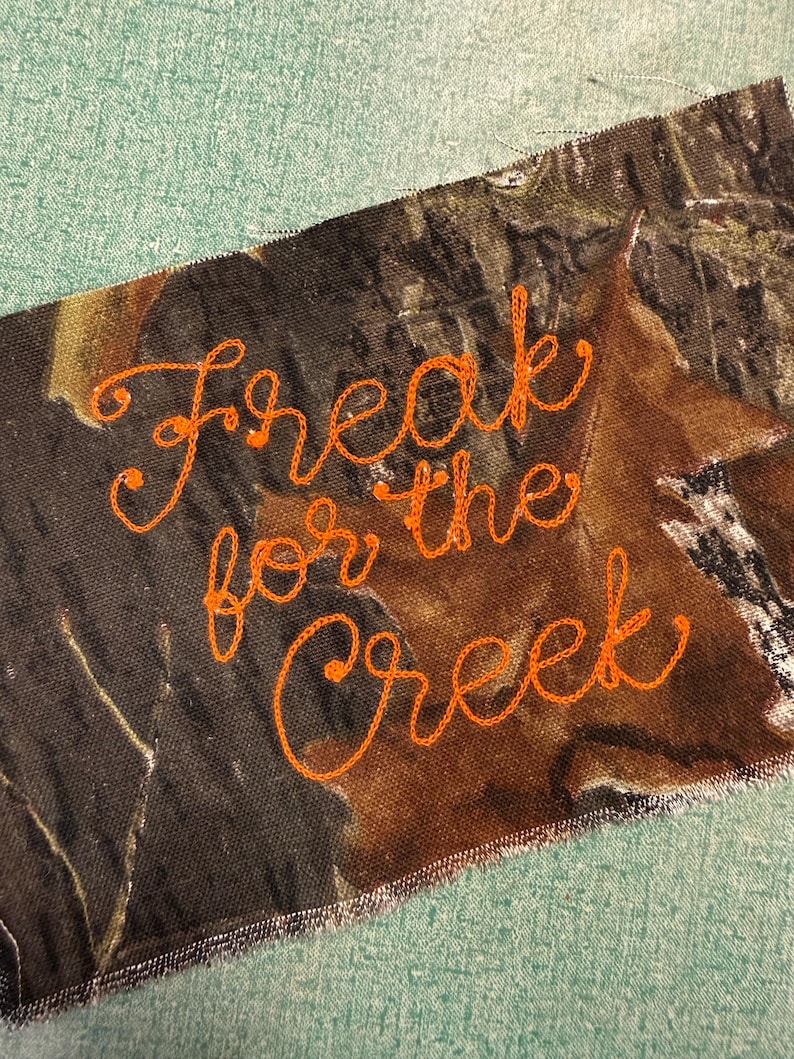 May include: A rectangular fabric patch featuring a camouflage pattern in shades of brown, black, and green. The text "Freak for the Creek" is embroidered in bright orange thread across the center in a cursive, handwritten-style script font.