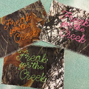 May include: Three rectangular fabric patches featuring a camouflage pattern with the embroidered text Freak for the Creek. One patch has orange stitching, one has bright pink stitching, and one has lime green stitching on the camo fabric.