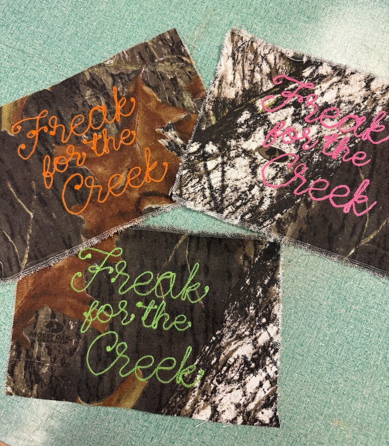 May include: Three rectangular fabric patches featuring a camouflage pattern with the embroidered text Freak for the Creek. One patch has orange stitching, one has bright pink stitching, and one has lime green stitching on the camo fabric.