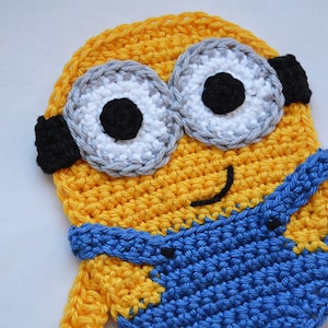 May include: A crocheted Minion character with yellow and blue yarn. The Minion has a big smile, two large eyes, and black goggles.