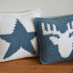 May include: Two square crochet cushions. One is white with a blue star design. The other is blue with a white moose head design. Both have a textured, chunky knit appearance.