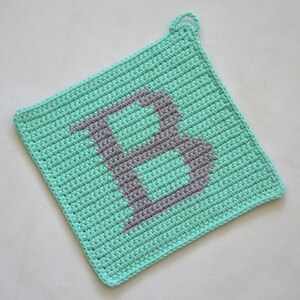 Könnte beinhalten: Ein türkisfarbener Topflappen aus Häkelarbeit mit einem grauen Buchstaben "B" in der Mitte.